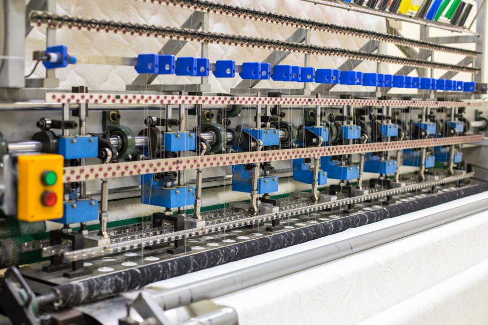 Automatyczna maszyna do pikowania tkanin z żółtym panelem sterowania z zielonym i czerwonym przyciskiem, pikująca białą tkaninę w regularny wzór.