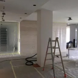 Przygotowanie do prac wykończeniowych w przestronnym wnętrzu: podłoga i drzwi zabezpieczone folią i taśmą, drabina ustawiona w centralnej części pomieszczenia, widoczne elementy instalacji...