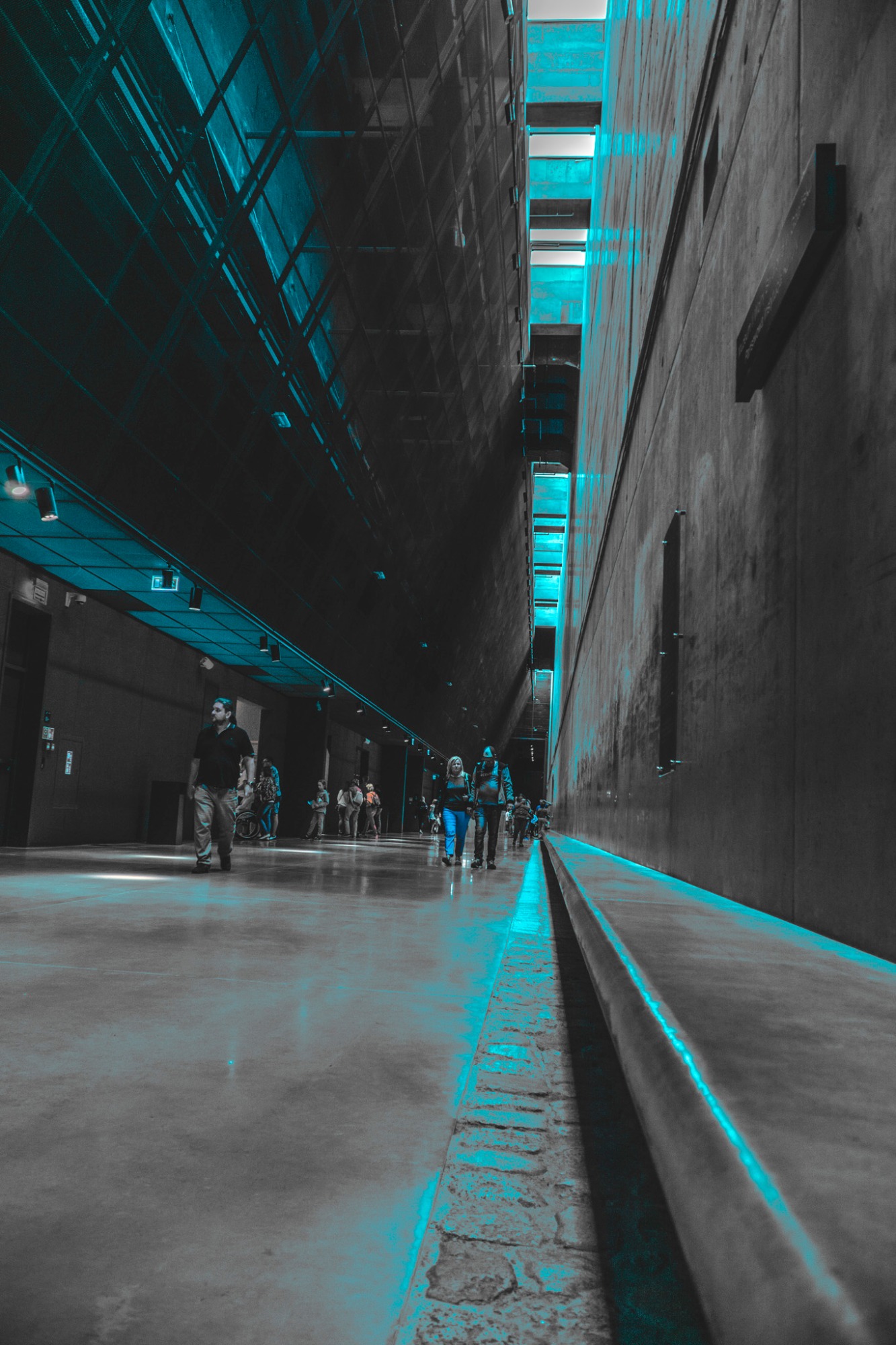 Wnętrze nowoczesnego budynku z wysokim sufitem, oświetlone niebieskim światłem, z przechodzącymi ludźmi. Betonowe ściany i podłoga w odcieniach szarości i niebieskiego.