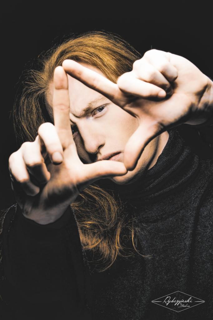 Portret młodego mężczyzny z rudymi włosami, w czarnym golfie, kadrującego twarz dłońmi na czarnym tle, z widocznym logo studia w dolnym rogu.