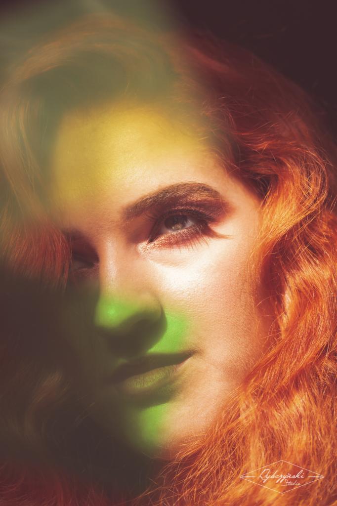 Portret kobiety z rudymi włosami, oświetlonej zielonym i żółtym światłem, z widocznym makijażem oczu i brwi.