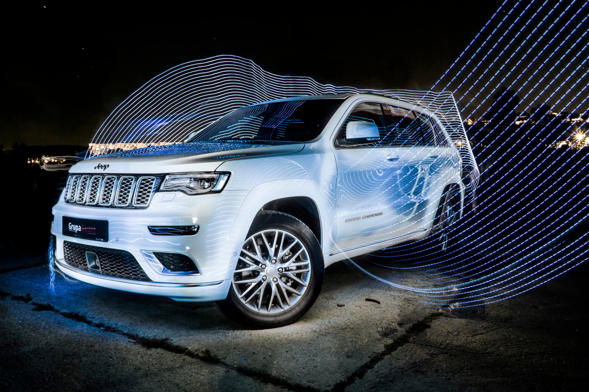 Biały Jeep Grand Cherokee sfotografowany w nocy z użyciem techniki light painting, tworzącej abstrakcyjne, niebieskie linie wokół samochodu, na tle rozświetlonego miasta.