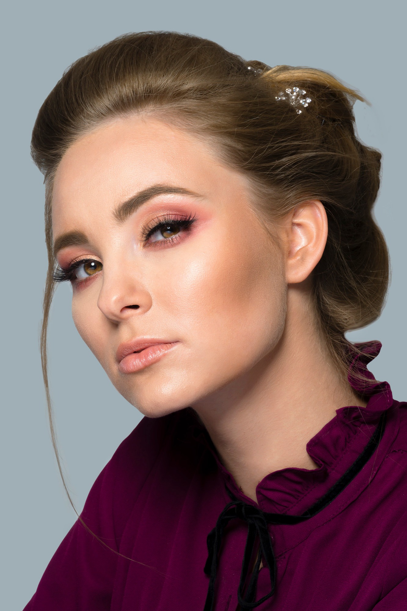 Portret kobiety z upiętymi włosami ozdobionymi błyszczącymi spinkami, w bordowej bluzce z wiązaniem przy szyi i z profesjonalnym makijażem oczu w odcieniach różu.