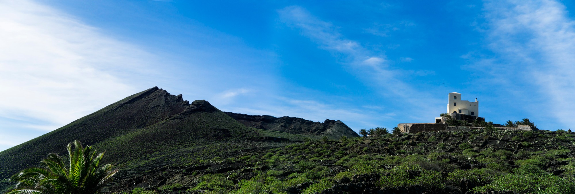 Panoramiczny widok na biały budynek z wieżą, położony na wzgórzu porośniętym zielenią, z wulkanicznym stożkiem w tle i błękitnym niebem z delikatnymi chmurami.