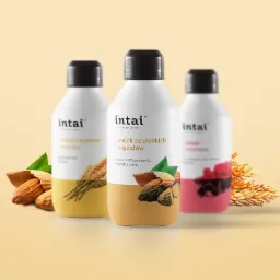 Trzy butelki olejków do ciała Intai: z kiełków pszenicy, ze słodkich migdałów i rycynowy, z elementami dekoracyjnymi z migdałów i pszenicy, na jasnym tle.