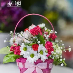 Różowo-biały koszyczek z mieszanką róż, margerytek i gipsówki, ozdobiony kokardą w kropki, z logo firmy w tle.