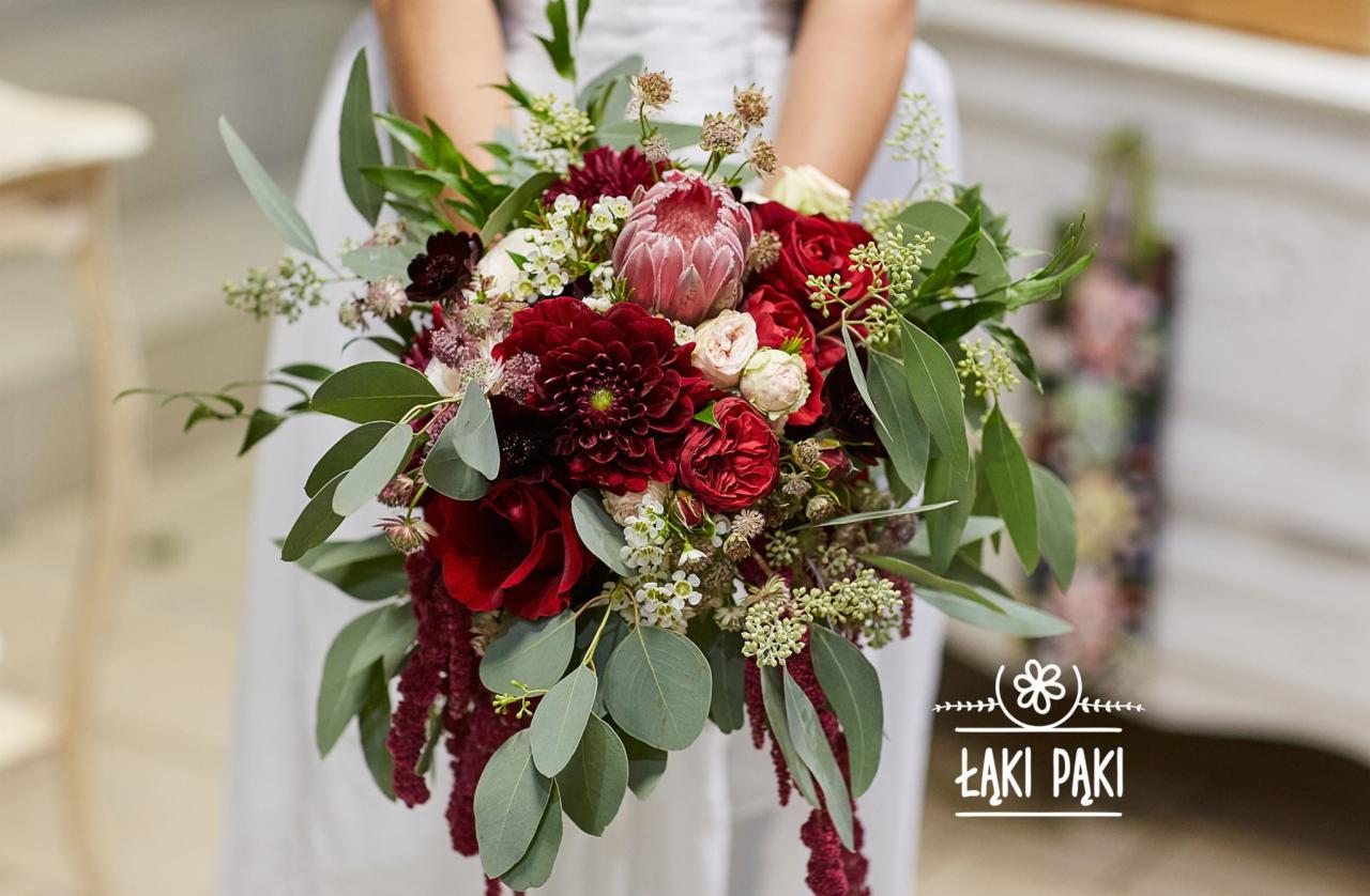 Kobieta w białej sukience trzyma bukiet z ciemnoczerwonych róż, dalii, protei królewskiej, eukaliptusa i drobnych białych kwiatów. Widoczne logo firmy florystycznej 'Łąki Pąki'.