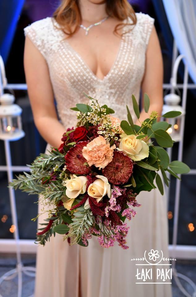 Panna młoda trzyma elegancki bukiet ślubny z róż, protei i eukaliptusa, z widocznym naszyjnikiem i białymi lampionami w tle.