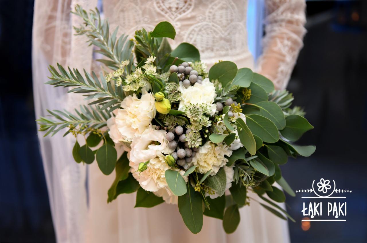 Panna młoda w koronkowej sukni trzyma bukiet ślubny z białych kwiatów, eukaliptusa i srebrnych jagód, z widocznym logo florysty na dole.