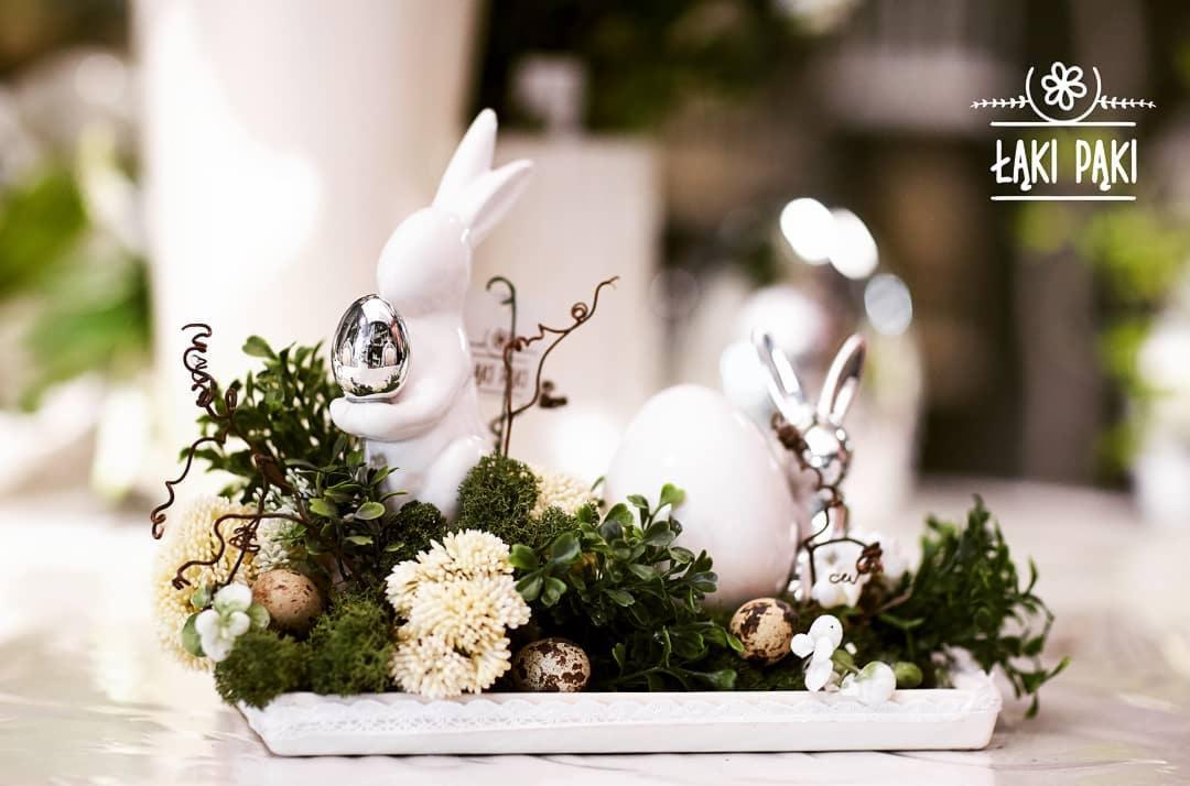 Dekoracja wielkanocna: biały ceramiczny królik trzymający srebrne jajko, kompozycja z zielonych roślin, mchu i przepiórczych jaj na białej tacy z koronkowym brzegiem, w tle logo 'Łąki Paki'.