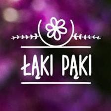 Logo firmy Łąki Pąki, przedstawiające stylizowany kwiat w okręgu z gałązkami po bokach, umieszczone nad napisem 'ŁĄKI PĄKI' na rozmytym tle w odcieniach fioletu.