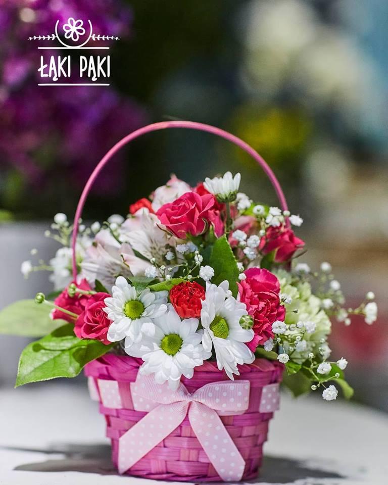 Różowo-biały koszyczek z mieszanką róż, margerytek i gipsówki, ozdobiony kokardą w kropki, z logo firmy w tle.