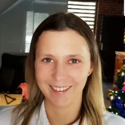 Uśmiechnięta kobieta w białej koszuli, z choinką w tle, prezentująca profesjonalny wizerunek.