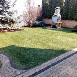 Perfekcyjnie utrzymany trawnik z wykoszonymi pasami, obok ścieżka z kostki brukowej i kamienny grill w tle, otoczony tujami.