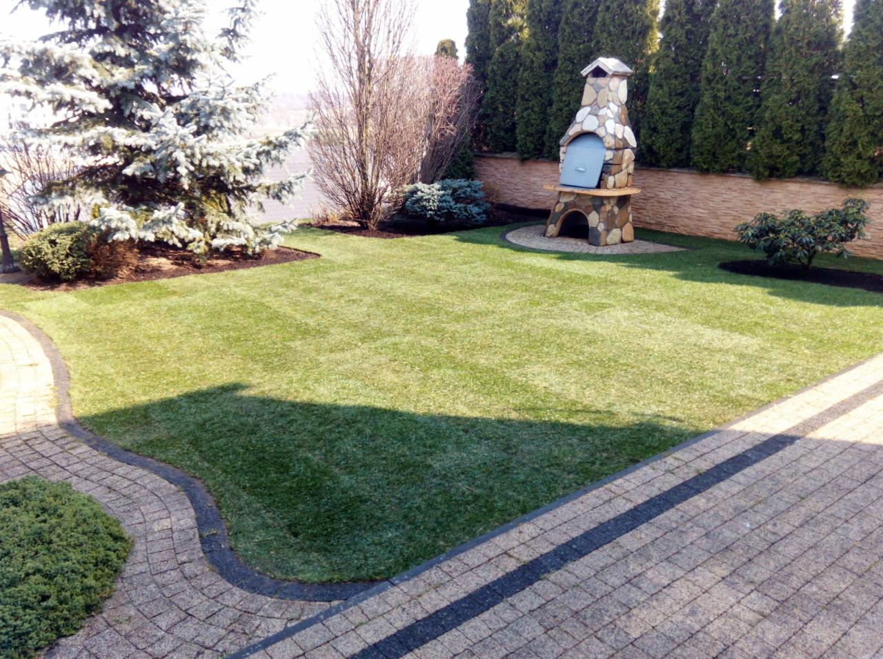 Perfekcyjnie utrzymany trawnik z wykoszonymi pasami, obok ścieżka z kostki brukowej i kamienny grill w tle, otoczony tujami.