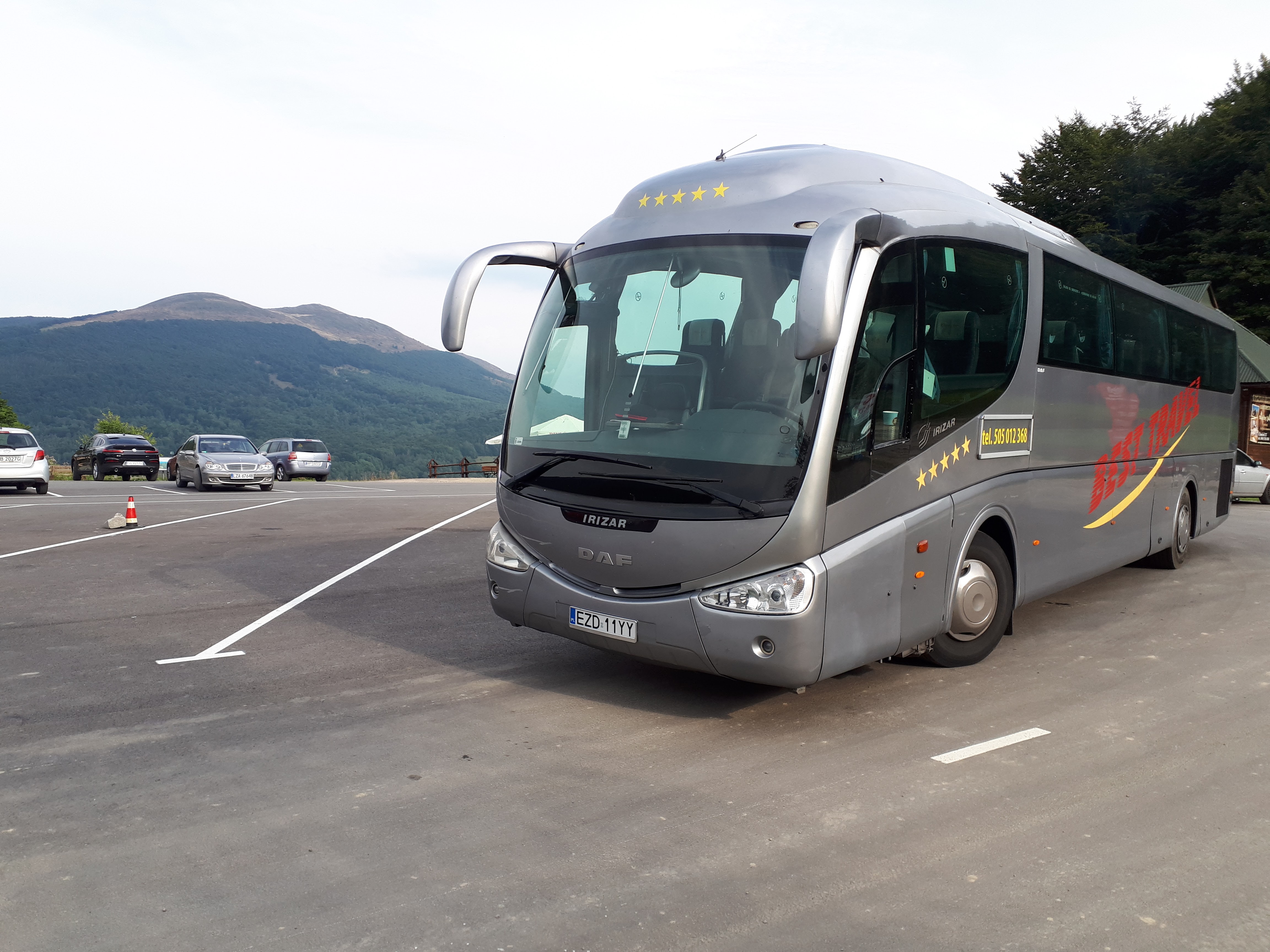 Szary autokar turystyczny marki DAF Irizar z pięcioma gwiazdami na dachu, zaparkowany na parkingu z widokiem na góry.