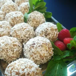 Srebrna taca z rzędami kokosowych kulek, dekorowanych świeżą miętą i malinami, na ciemnoniebieskim tle.