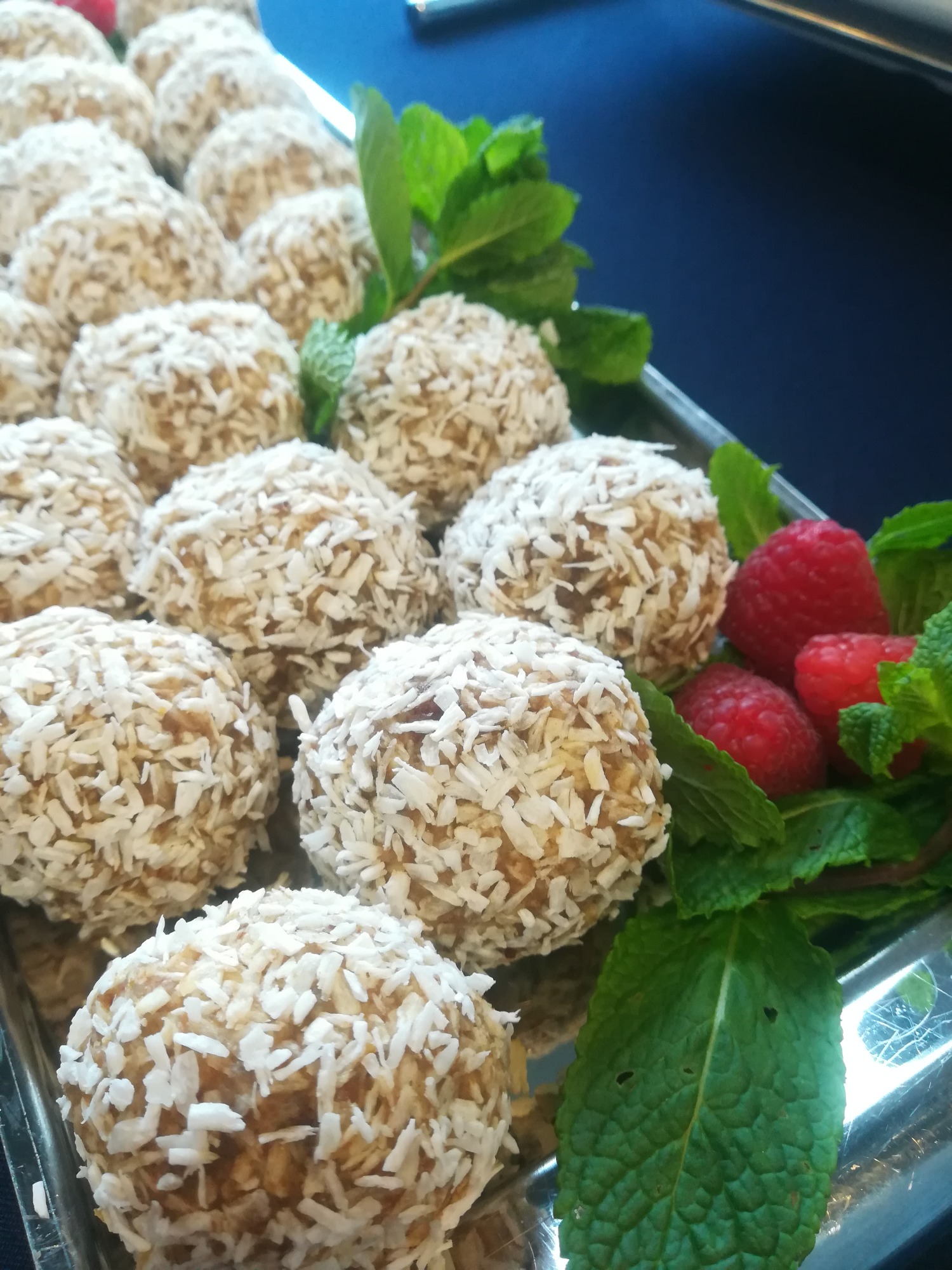 Srebrna taca z rzędami kokosowych kulek, dekorowanych świeżą miętą i malinami, na ciemnoniebieskim tle.