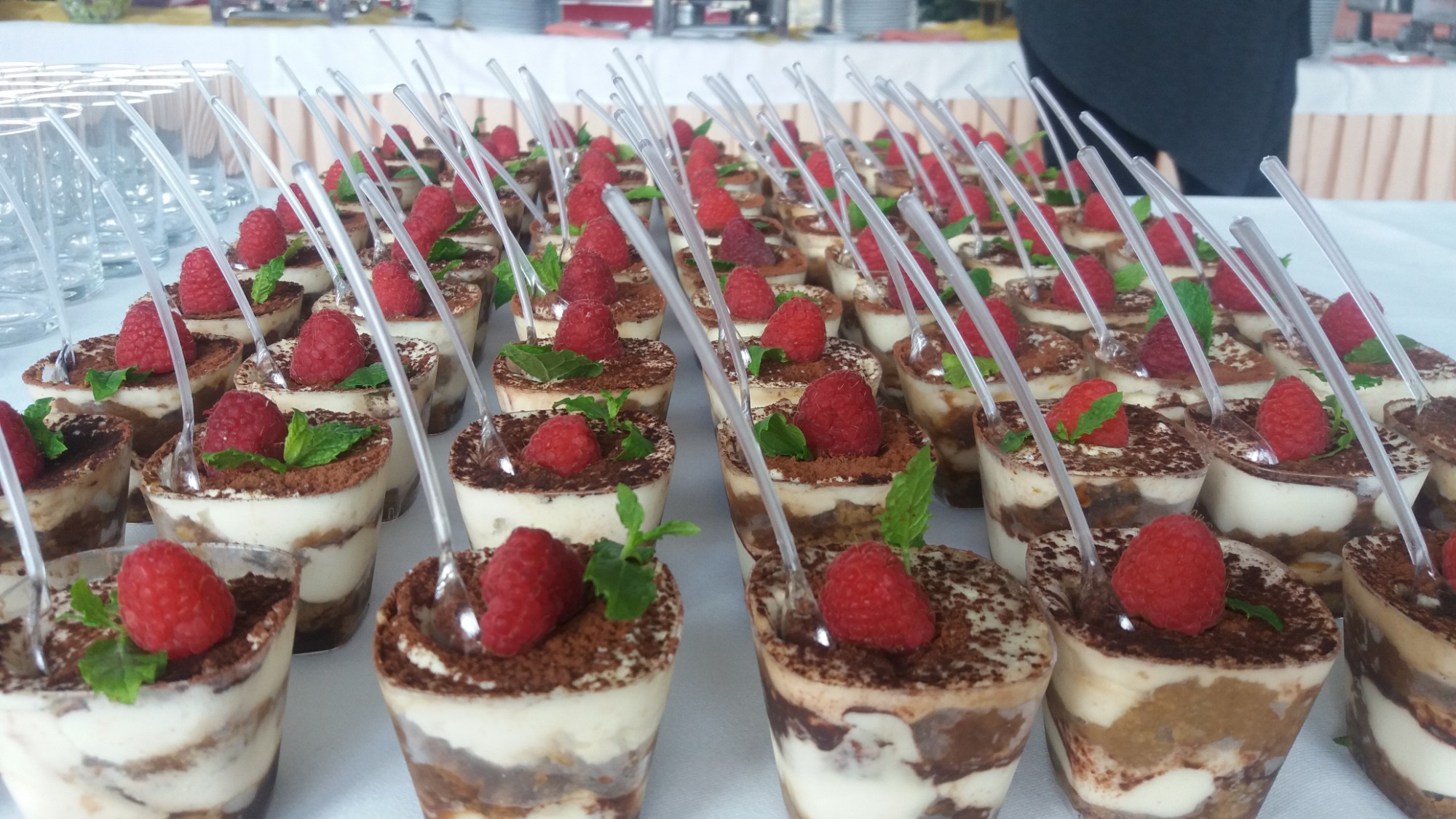 Rząd deserów tiramisu w plastikowych kubeczkach, każdy udekorowany maliną i listkiem mięty, z plastikową łyżeczką wbita w środek, ułożonych na białym stole.