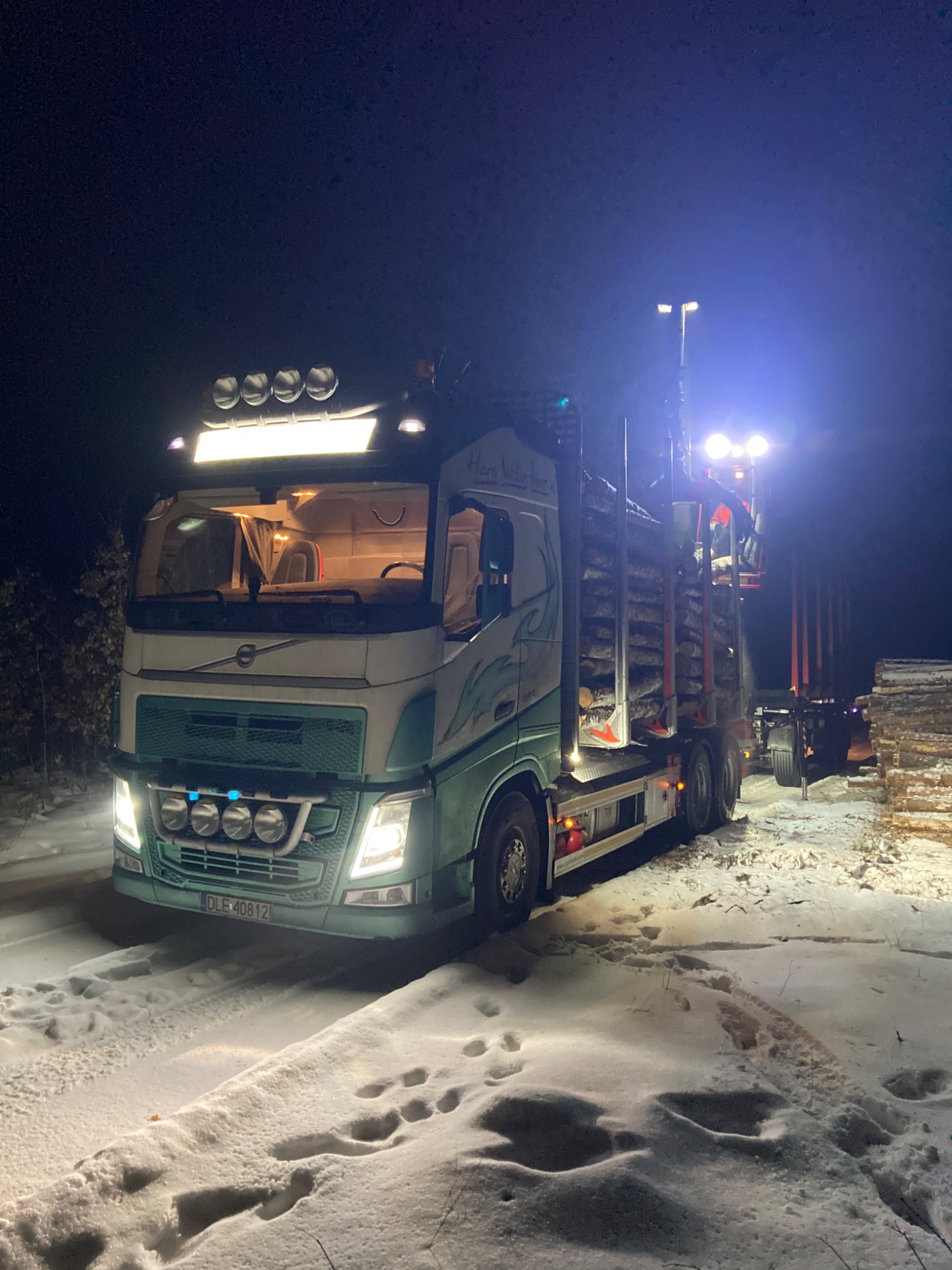 Ciężarówka Volvo FH załadowana drewnem, oświetlona reflektorami, stoi na ośnieżonej drodze w nocy, widoczne ślady opon w śniegu i tablica rejestracyjna DLE 40812.