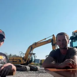 Dwóch mężczyzn pozuje na tle żółtej koparki i ciężarówki Scania, na żwirowym terenie pod błękitnym niebem. Jeden w czapce i okularach, drugi oparty o blat.