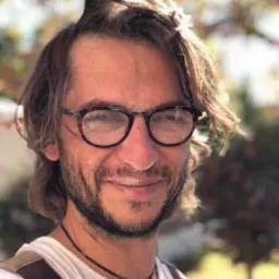 Uśmiechnięty mężczyzna w okularach, z mąką na nosie i twarzy, włosy częściowo związane, na zewnątrz, w tle drzewa.