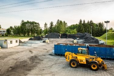 Żółta ładowarka JCB na placu składowym z hałdami czarnego węgla i niebieskimi kontenerami, w tle budynek i drzewa.