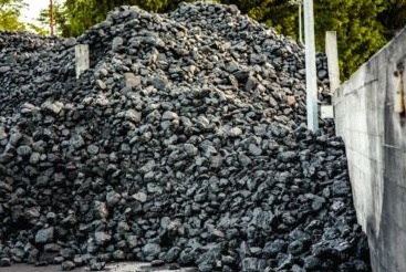 Góra antracytu o nieregularnych kształtach, składowana przy betonowym murze z widocznymi śladami użytkowania, z zielenią drzew w tle.