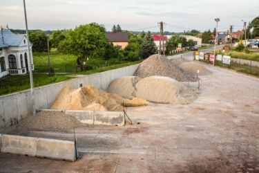 Składowisko różnych frakcji węgla i kruszyw, widoczne pryzmy piasku, żwiru i węgla na betonowym placu, w tle zabudowania wiejskie i zieleń.
