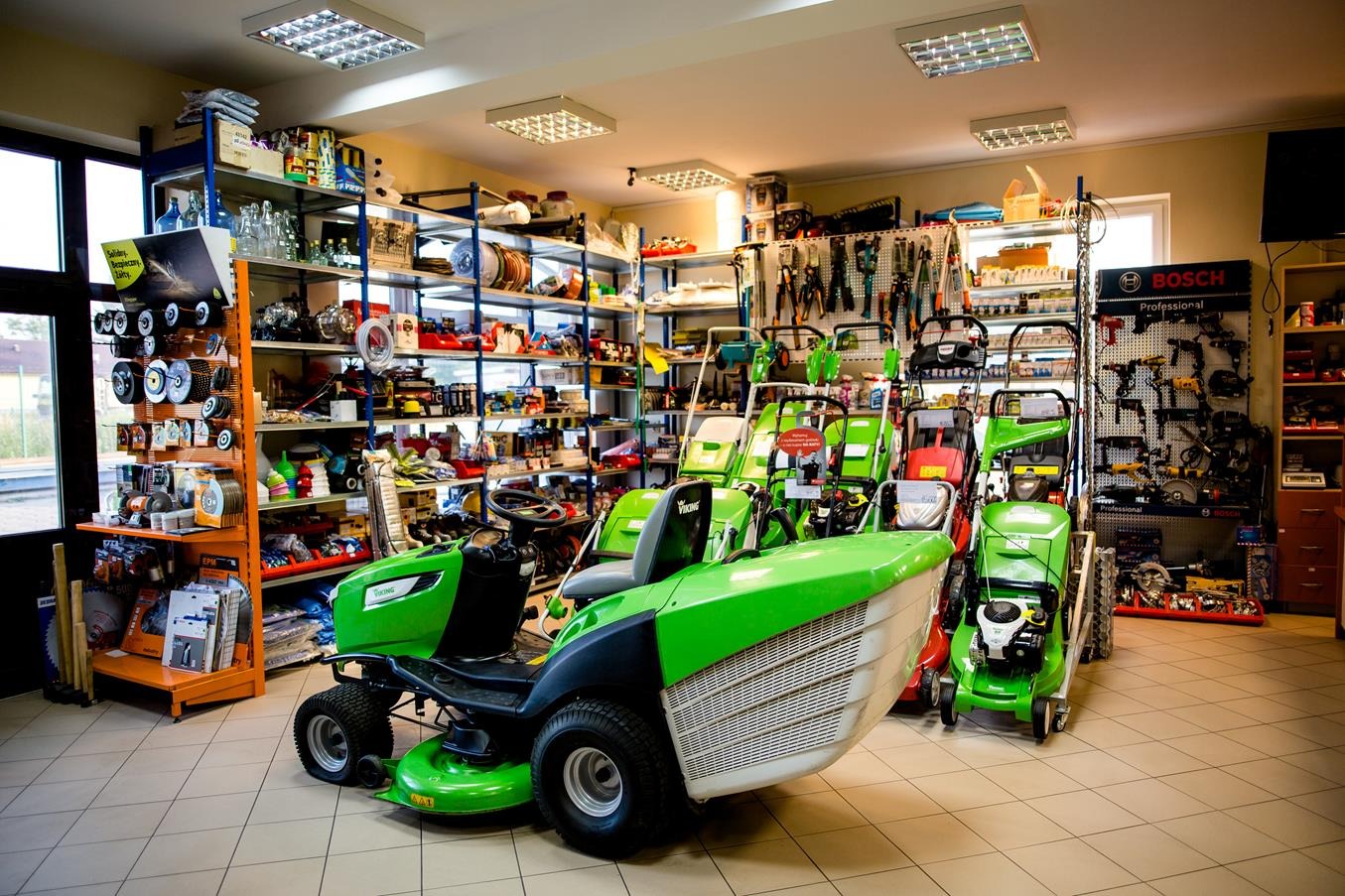 Wnętrze sklepu z kosiarkami do trawy, w tym kosiarki traktorowe i tradycyjne, narzędzia ogrodnicze na półkach, oświetlenie sufitowe.