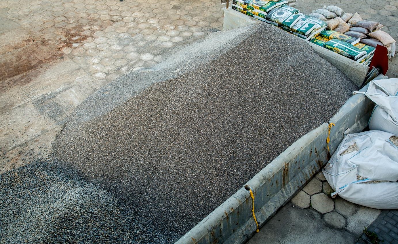 Góra drobnego, szarego kruszywa w betonowym boksie, obok worki z materiałem i palety z zapakowanym towarem, widok z góry.