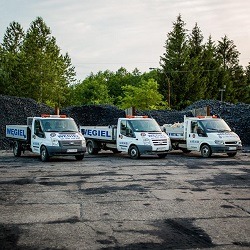 Trzy identyczne samochody dostawcze z napisem 'WĘGIEL' na boku, ustawione rzędem na placu, w tle hałda węgla i drzewa.
