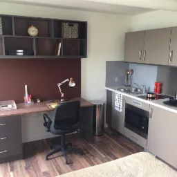 Wnętrze kawalerki z aneksem kuchennym i biurkiem z szufladami oraz półkami wiszącymi, utrzymane w szaro-brązowej kolorystyce z drewnianą podłogą.