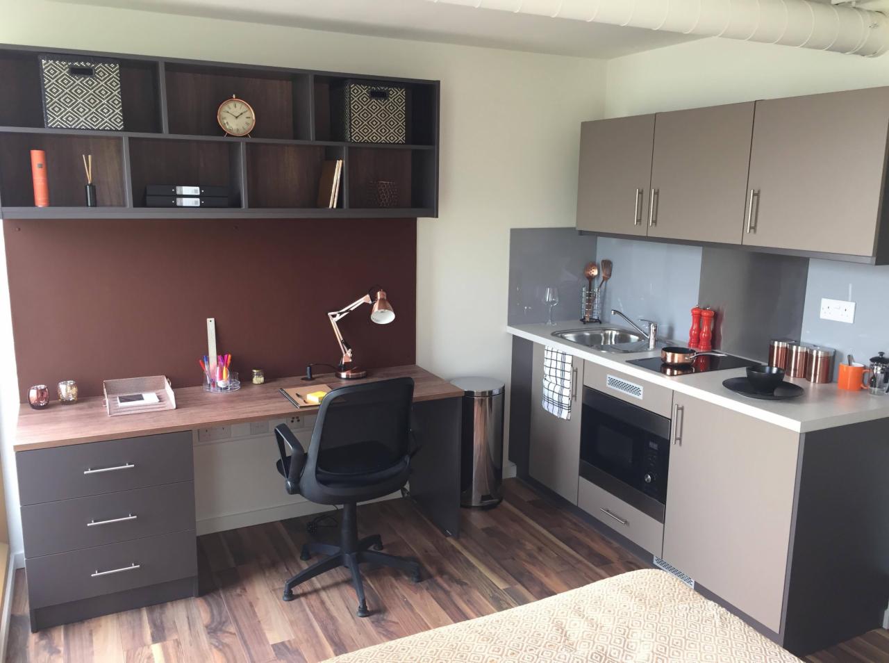 Wnętrze kawalerki z aneksem kuchennym i biurkiem z szufladami oraz półkami wiszącymi, utrzymane w szaro-brązowej kolorystyce z drewnianą podłogą.