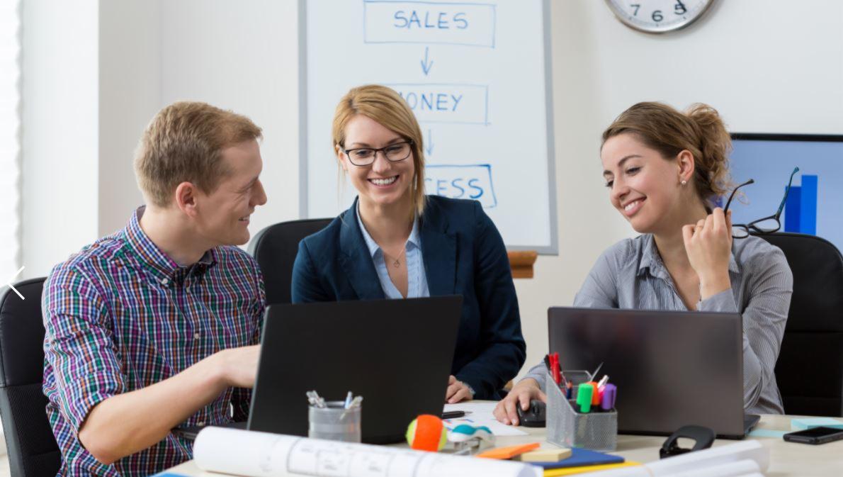 Trzy osoby uśmiechnięte, pracujące przy laptopach w biurze; na tablicy schemat blokowy z hasłami: Sales, Money, Success; na biurku dokumenty i przybory biurowe.