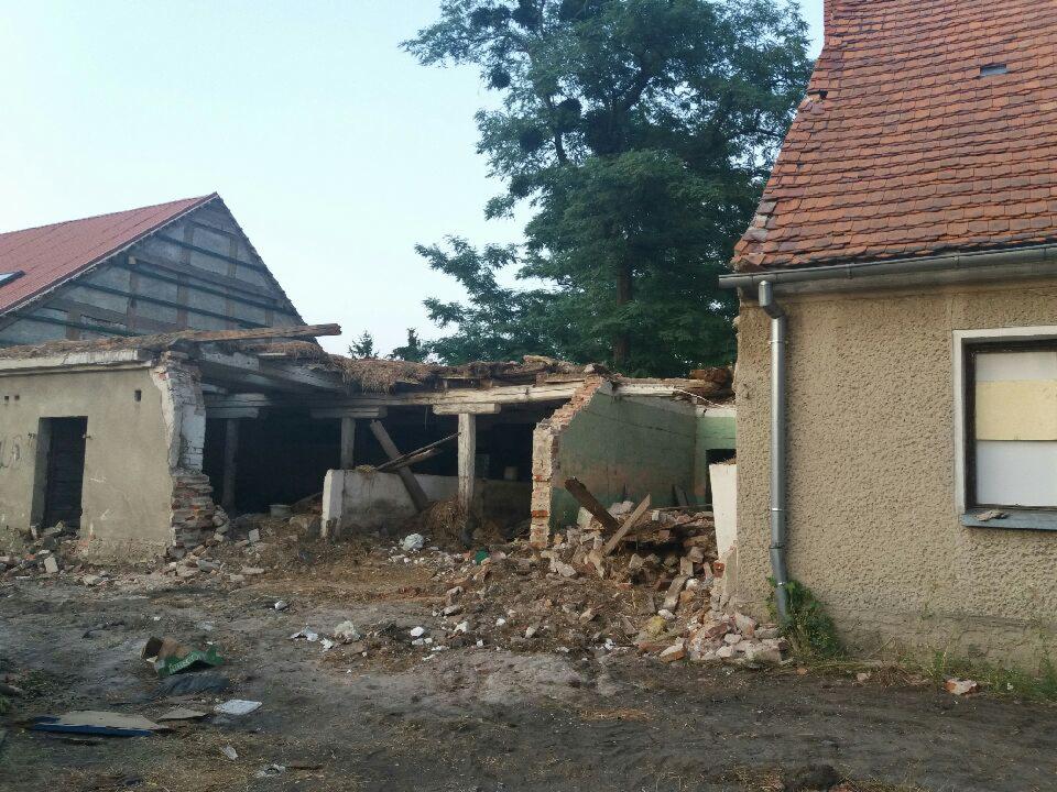 Częściowo rozebrany budynek z cegły i drewna, gruz i błoto na ziemi, widoczne fragmenty dachu i ścian sąsiednich budynków, zielone drzewo w tle.