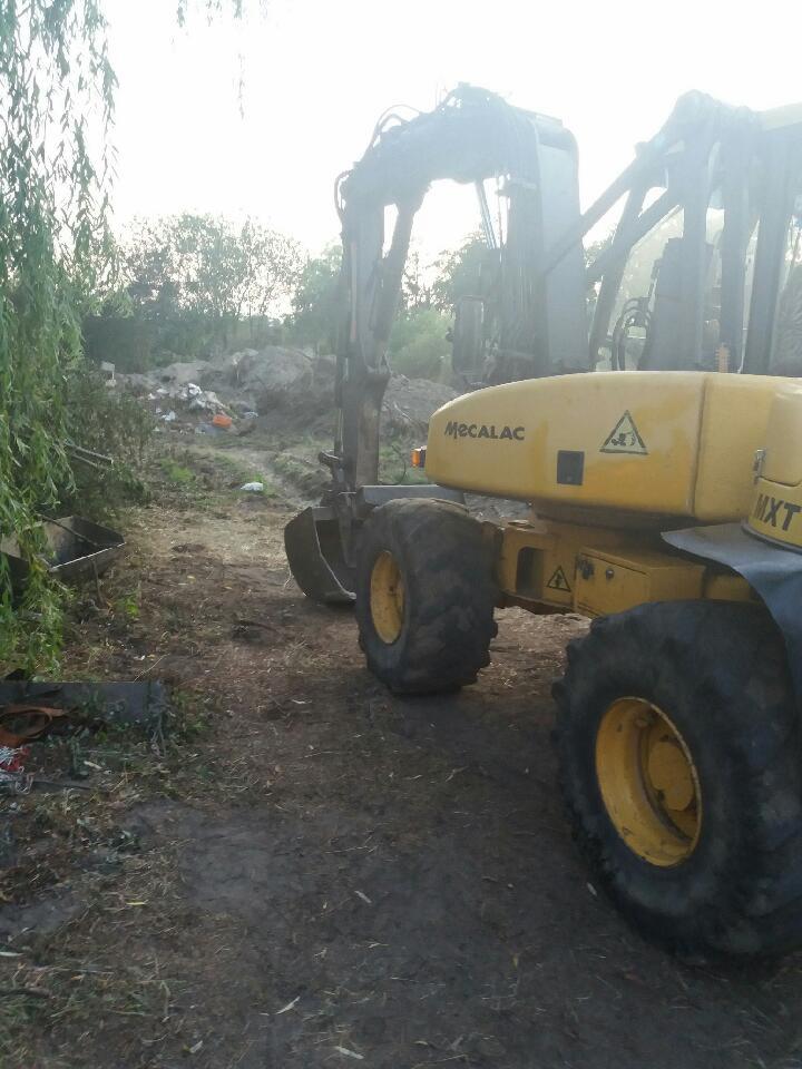 Żółta koparka Mecalac MXT z łyżką na błotnistym terenie, obok drzewa i śmieci, w pochmurny dzień. Widoczne duże opony i ramię koparki.