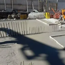 Wylewanie betonu na przygotowany szalunek z wystającymi prętami zbrojeniowymi, widoczne narzędzia i sprzęt budowlany w tle.