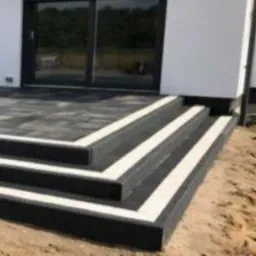 Schody i taras z ciemnej kostki brukowej z białymi fugami prowadzące do nowoczesnego domu z czarnymi ramami okiennymi i drzwiami tarasowymi, widok na plac budowy.