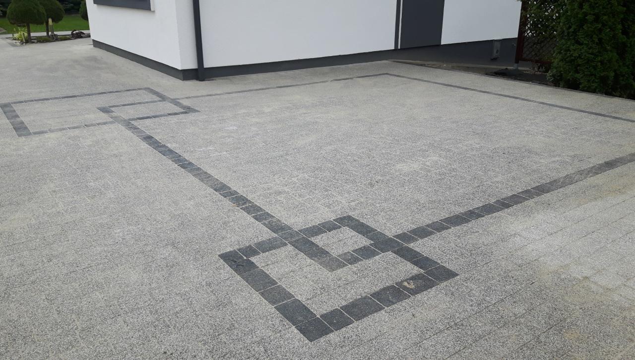 Ułożona jasnoszara kostka brukowa z ciemnoszarą obwódką tworzącą geometryczny wzór przed białym budynkiem z ciemnoszarą podmurówką.