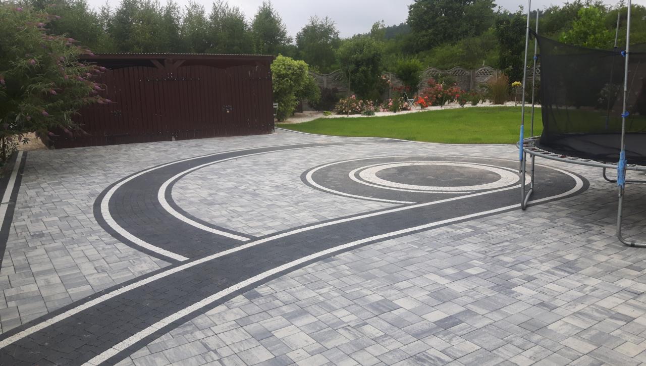 Ułożona kostka brukowa na podjeździe z dekoracyjnymi, ciemnymi pasami i okręgiem, w tle drewniany garaż i trampolina.