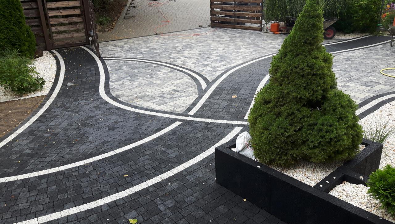 Ułożona nawierzchnia z kostki brukowej w dwóch odcieniach szarości, z białymi pasami oddzielającymi strefy, prowadząca do drewnianej bramy w tle, z ozdobnym drzewkiem iglastym w czarnej donicy po...