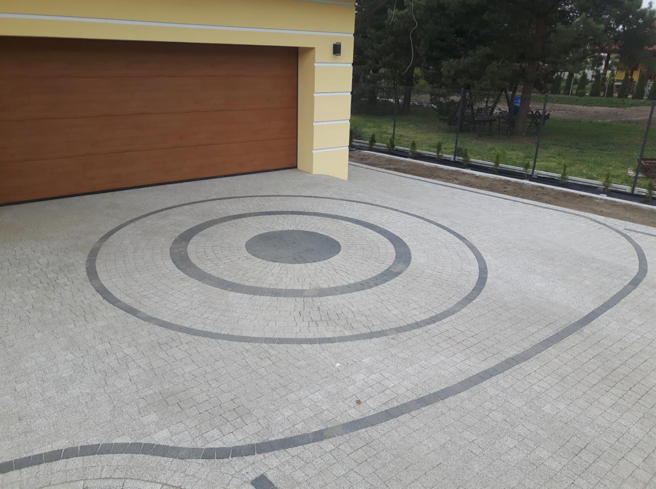 Podjazd do garażu wyłożony jasnoszarą kostką brukową z dekoracyjnym wzorem koncentrycznych okręgów i falistą linią z ciemniejszej kostki.
