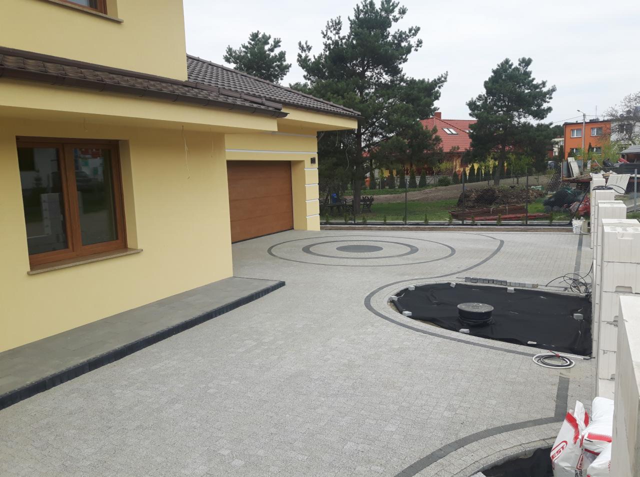 Ułożona szara kostka brukowa z dekoracyjnymi okręgami i obrzeżami przy żółtym domu z brązową bramą garażową; widoczny częściowo przygotowany teren z czarną folią, prawdopodobnie pod oczko wodne...