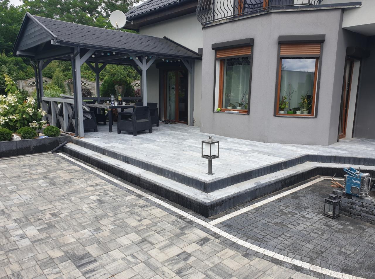 Nowoczesny taras z szarej kostki brukowej prowadzący do altany ogrodowej, obrzeża z ciemnej kostki, widoczny fragment elewacji domu z oknami i lampą ogrodową.