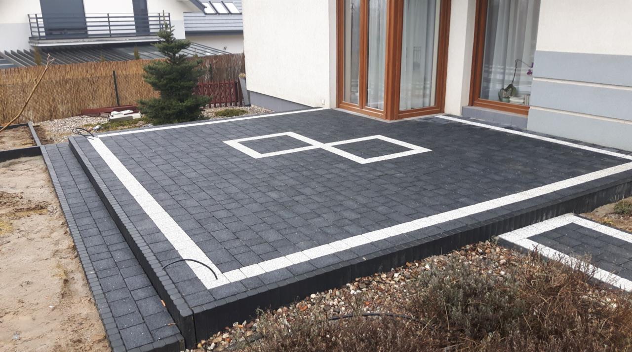 Nowoczesny taras z ciemnej kostki brukowej z białymi akcentami w kształcie dwóch kwadratów, schody z kostki prowadzące na trawnik, widok na dom z oknami.