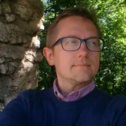 Mężczyzna w okularach, w niebieskim swetrze, pozuje na tle zieleni i kamiennej konstrukcji. Uśmiech na twarzy, naturalne światło, bliski kadr.