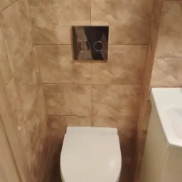 Wykończona łazienka z beżowymi płytkami imitującymi marmur, biała toaleta podwieszana z chromowanym przyciskiem spłukującym i fragmentem białej umywalki.