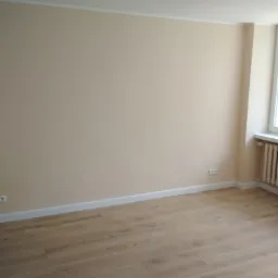 Pomalowane na beżowo wnętrze pokoju z panelami podłogowymi o jasnym odcieniu drewna, widoczne gniazdka elektryczne i białe listwy przypodłogowe.