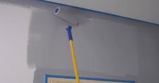 Wałek z niebieską rączką maluje szarą ścianę, zabezpieczoną niebieską taśmą malarską przy suficie i ościeżnicy.